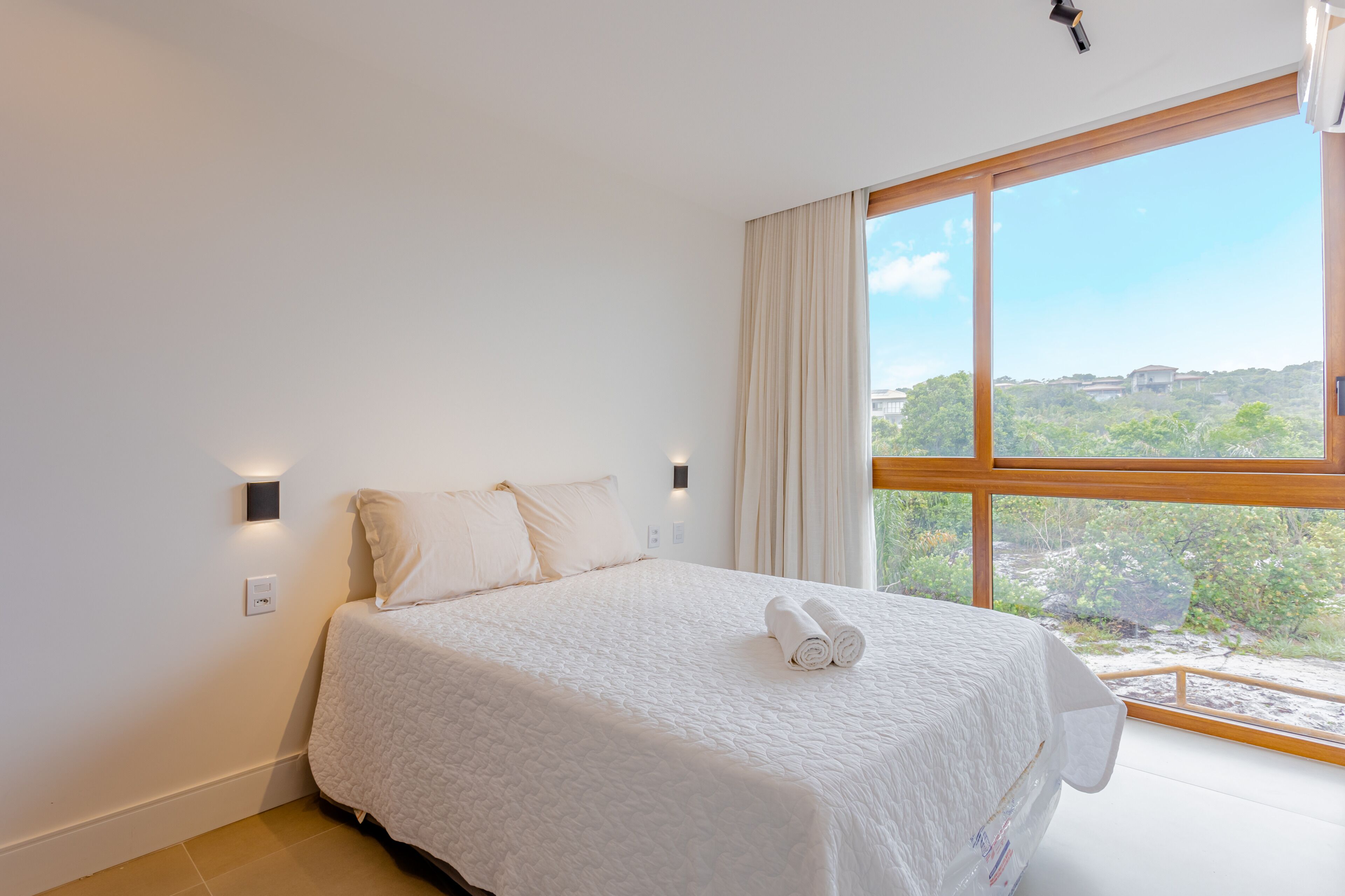 Ein ordentlich bezogenes Bett mit weißen Leinen und einem zusammengerollten Handtuch, ein großes Fenster mit Vorhängen und Blick ins Grüne.