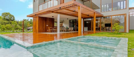 Una casa moderna con una piscina grande, terraza de madera y un espacio exterior cubierto.