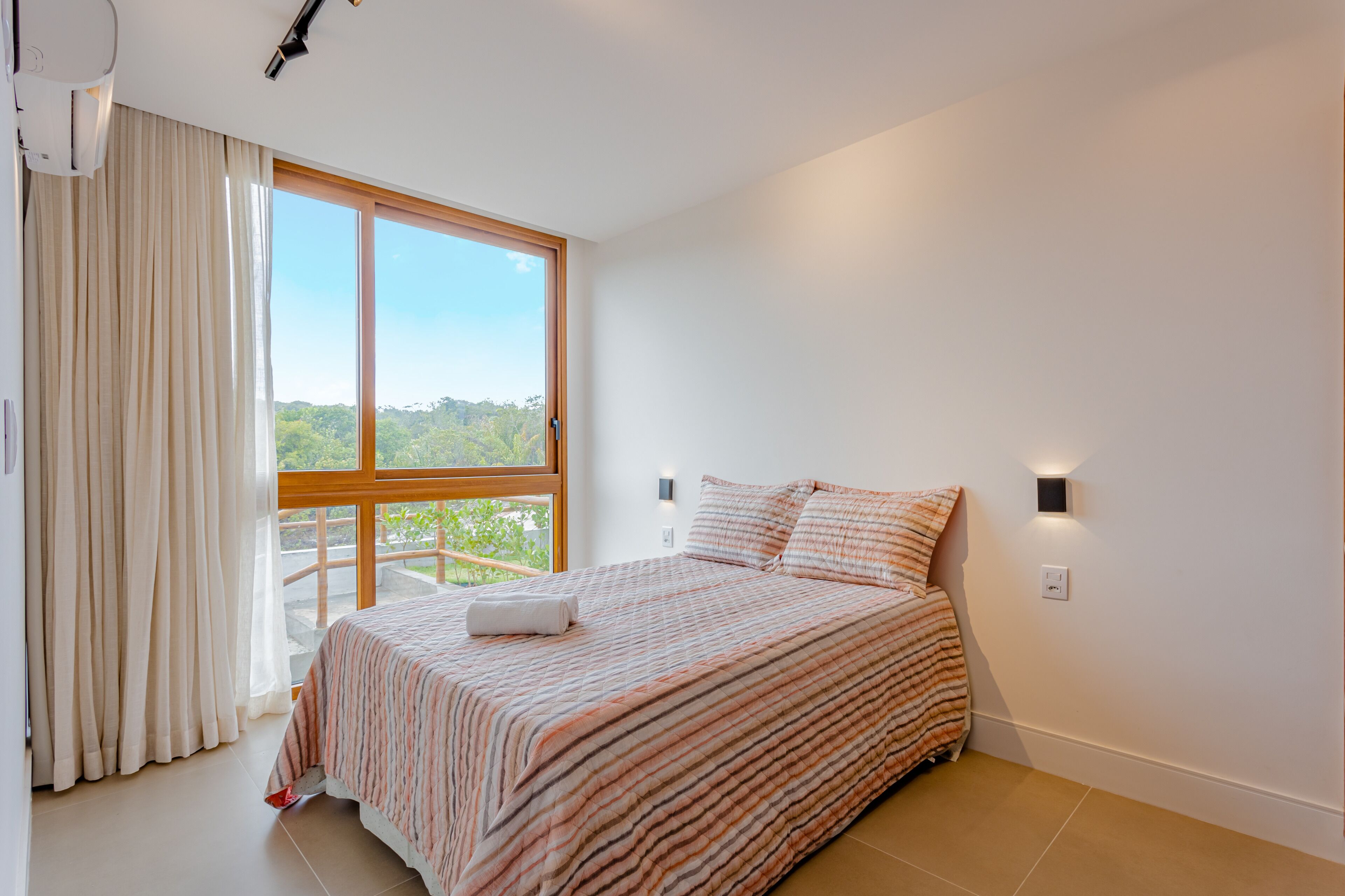 Une chambre avec une grande fenêtre, un lit avec une literie à rayures, un rideau et une lampe fixée au mur.
