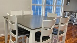 Un comedor con una mesa redonda y sillas blancas, ventanas amplias con persianas, y suelo de madera.