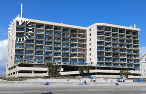 Un hotel de varios pisos con balcones, un aparcamiento y una zona de playa con sombrillas.