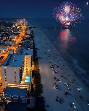 A coastal cityscape at night with a beach, buildings, and fireworks.