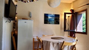 Une cuisine de taille modeste, avec un réfrigérateur blanc, des chaises en bois, une table recouverte d’une nappe, un luminaire suspendu et un tableau encadré accroché au mur.