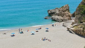Ein Strand mit klarem, blauem Wasser, Felsformationen und Menschen, die unter Sonnenschirmen entspannen.