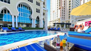 Una piscina con sillones reclinables azules y una mesa con bebidas, rodeada de altos edificios con ventanales arqueados.
