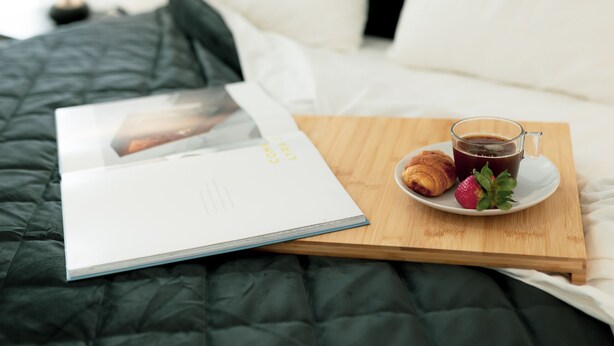 Ein ordentlich gemachtes Bett mit einer dunkelgrünen Decke, ein Holztischchen mit einer Tasse Kaffee, einem Croissant und einer Erdbeere auf einem weißen Teller, und eine aufgeschlagene Zeitschrift auf dem Bett.