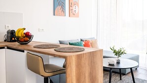 A modern kitchen with a wooden countertop, a bowl of fruit, and a coffee machine. There is a dining area with a chair and a small table. The room has a sofa and a plant on a side table. - Pebble Garden (Kos)