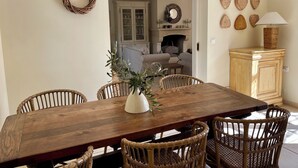 A dining area with a wooden table and wicker chairs, a white vase with greenery, and a wooden cabinet with a lamp.