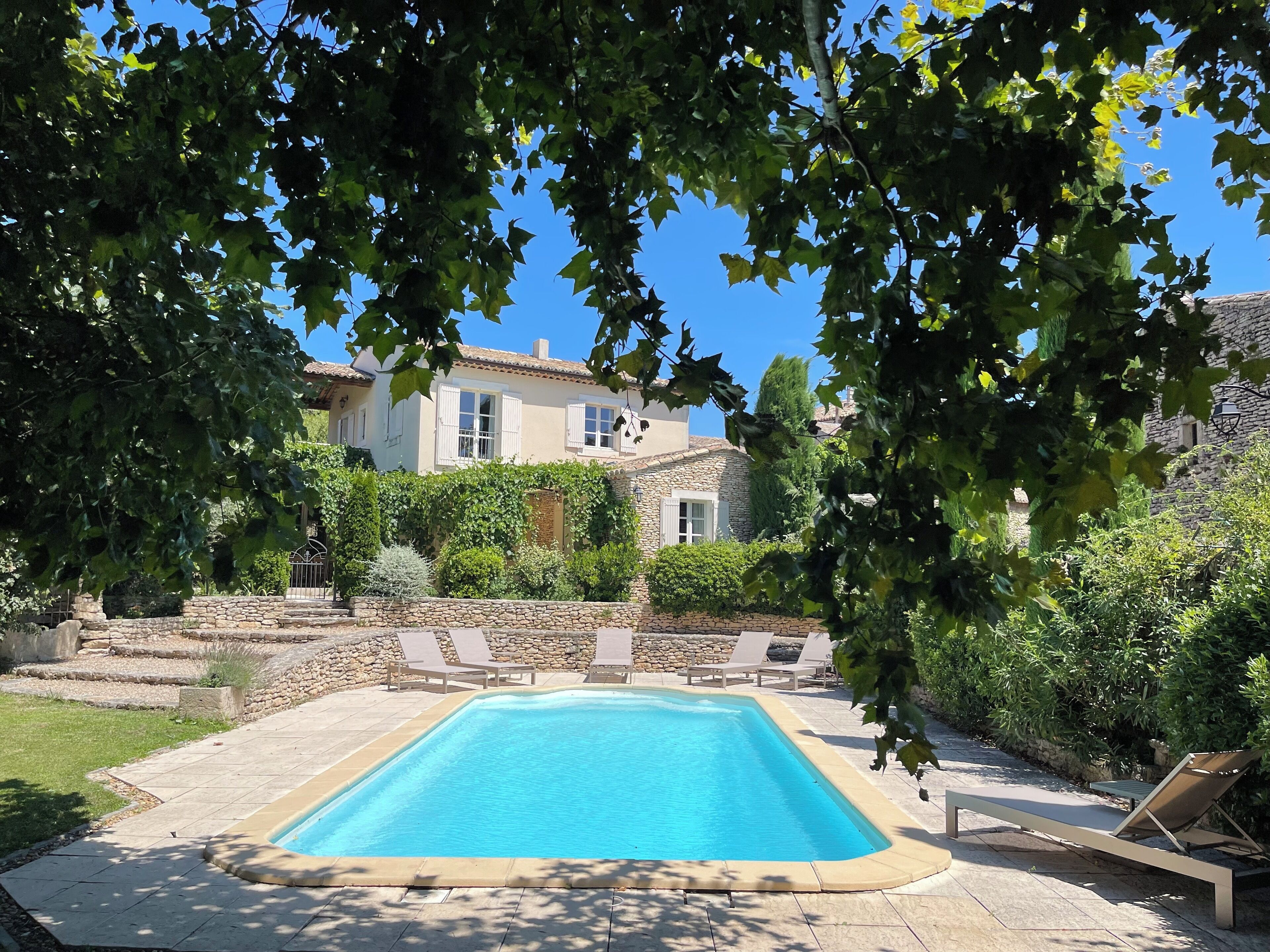 A swimming pool with lounge chairs, surrounded by a stone wall and greenery, with a house in the background.