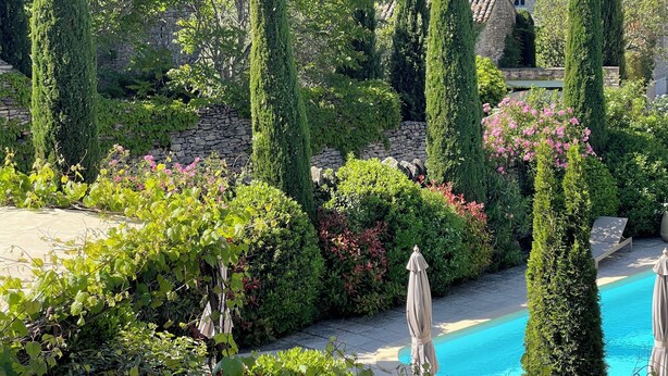 A landscaped garden with a swimming pool, tall cypress trees, and a stone wall.