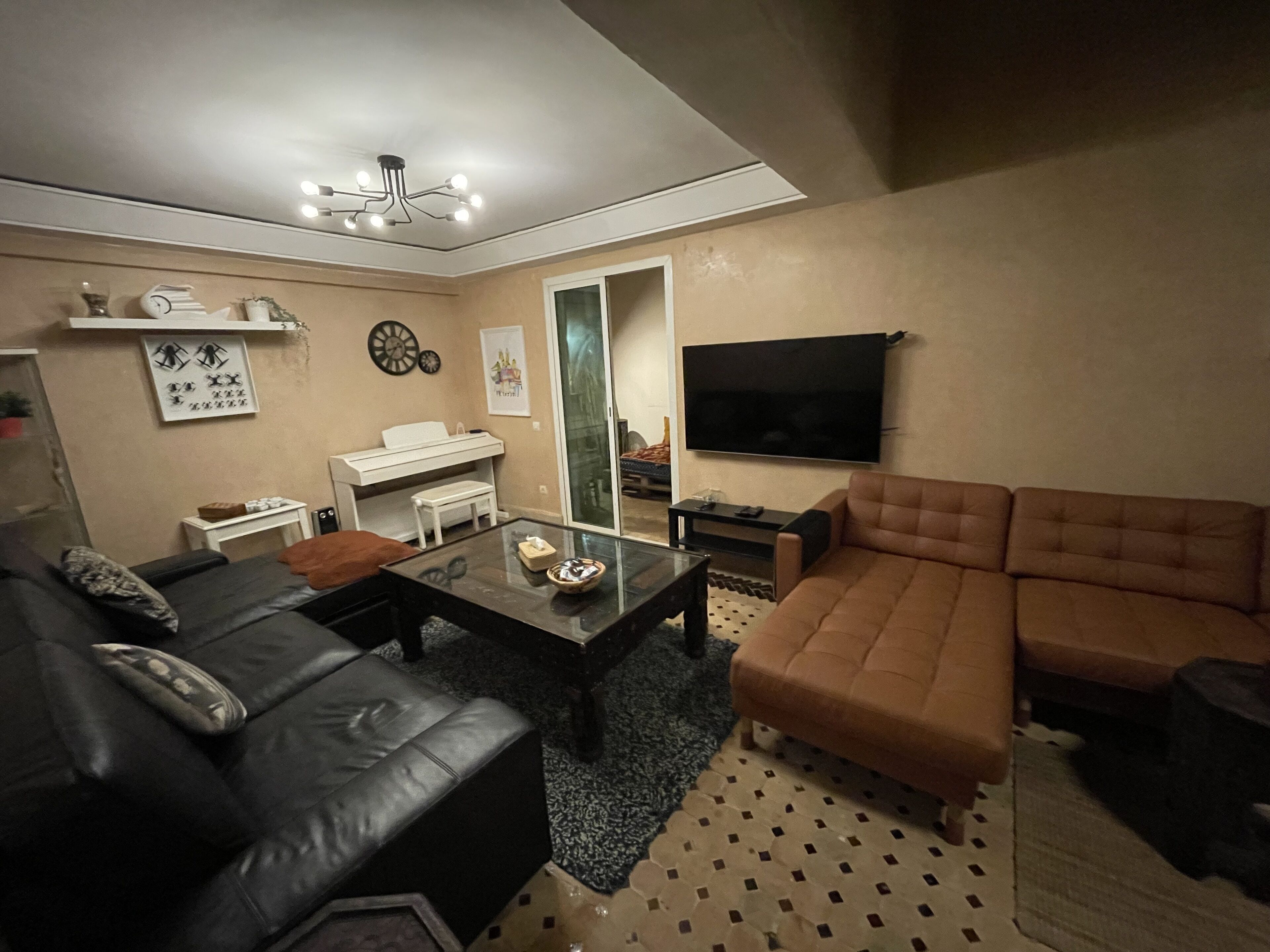 A living room with a black leather sofa, a brown leather couch, a glass coffee table, a white piano, a flat-screen TV, and a patterned rug.