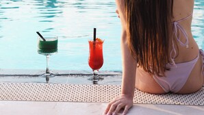 A pool area with a person sitting by the pool, two drinks on the edge, and palm trees in the background. - Luxury Premium Finese Hotel & SPA (Golem)