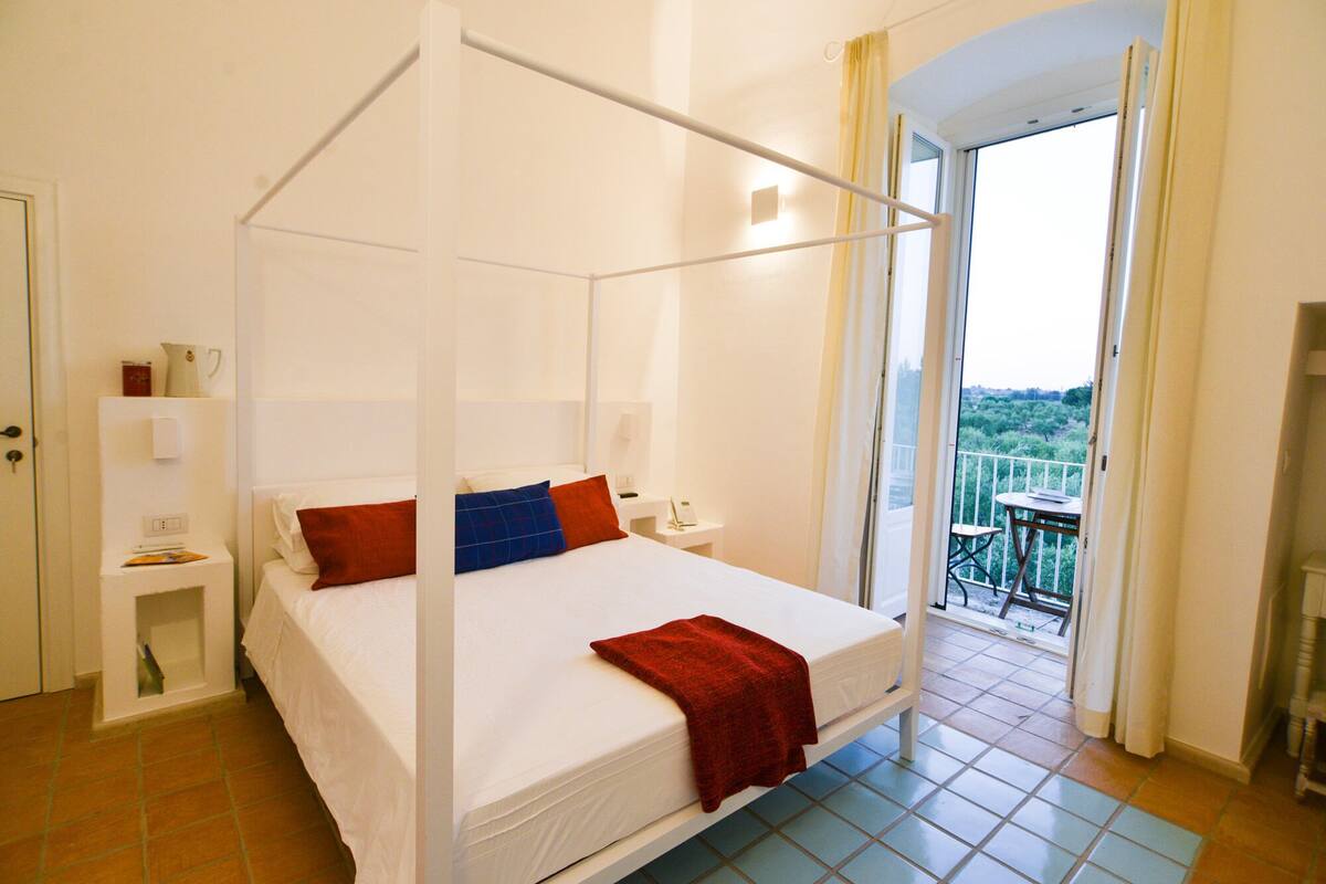 A four-poster bed with white linens and red and blue pillows, a bedside table with a lamp, and a view of a balcony with a table and chairs.