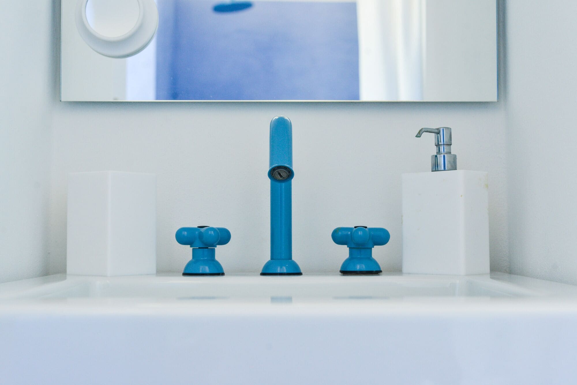 Un baño con un lavamanos blanco, un grifo azul y un dispensador de jabón blanco.