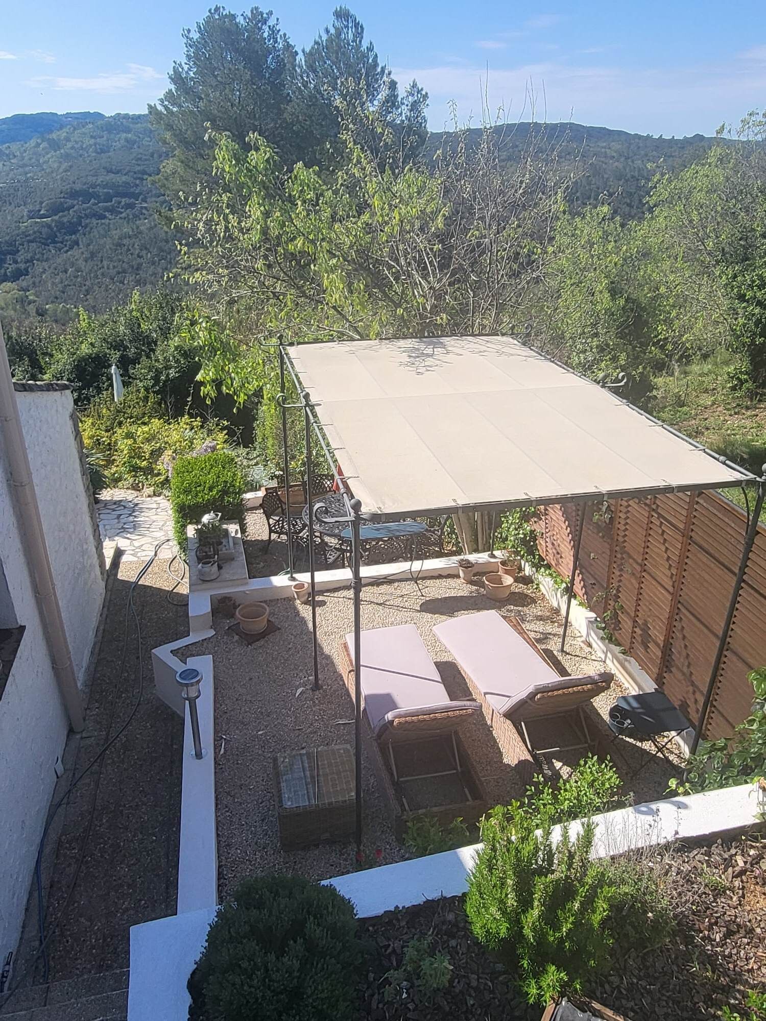 Un patio con zona de estar techada, una mesa y sillas. Hay plantas en macetas y un pequeño jardín.