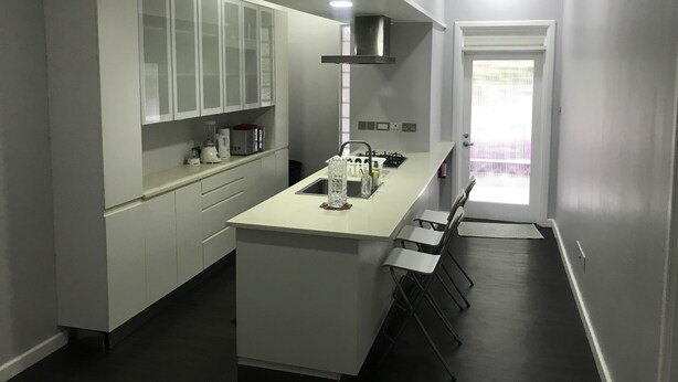 Une cuisine moderne avec des meubles blancs, un îlot central doté de tabourets, une cuisinière encastrée et une fenêtre avec des stores.