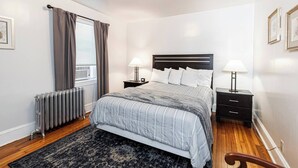 A bedroom with a bed, bedside tables, a radiator, a window with curtains, and a wooden floor. - Bryn Mawr Has It All (Bryn Mawr)
