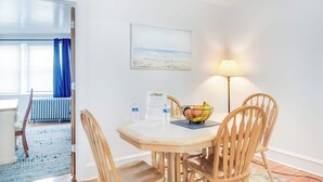 A dining area with a table, chairs, a bowl of fruit, a lamp, and a window with a view of the beach.