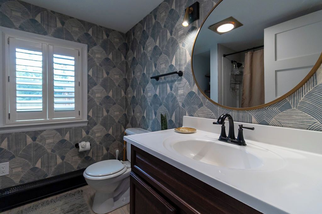Un bagno con lavandino bianco, mobile bagno in legno scuro, specchio rotondo e carta da parati decorata.