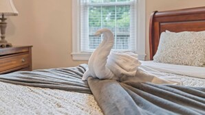 A neatly made bed with a swan-shaped towel, a wooden nightstand with a lamp, and a window with blinds. - Stay At Our Tasteful, Private, & Elegant Abode (Devon)