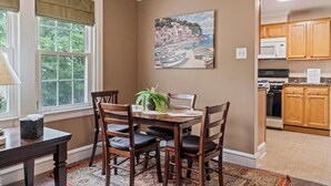 A dining area with a wooden table and chairs, a painting of a coastal scene, and a kitchen with wooden cabinets and appliances. - Stay At Our Tasteful, Private, & Elegant Abode (Devon)