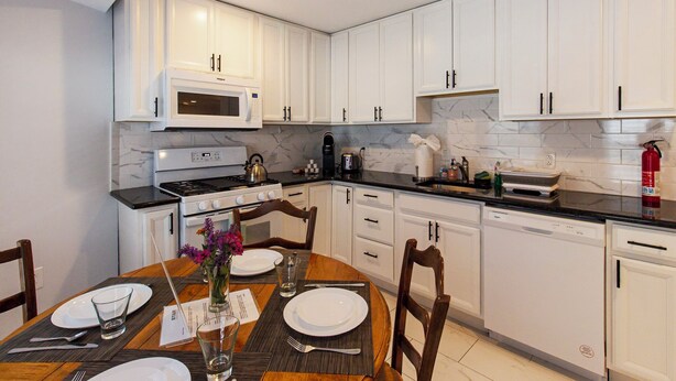 A modern kitchen with white cabinets, a built-in microwave, stove, and dishwasher. There is a dining table set with plates, glasses, and cutlery. - Devon - Horse Show, History, Walk to Shops, Café's (Devon)