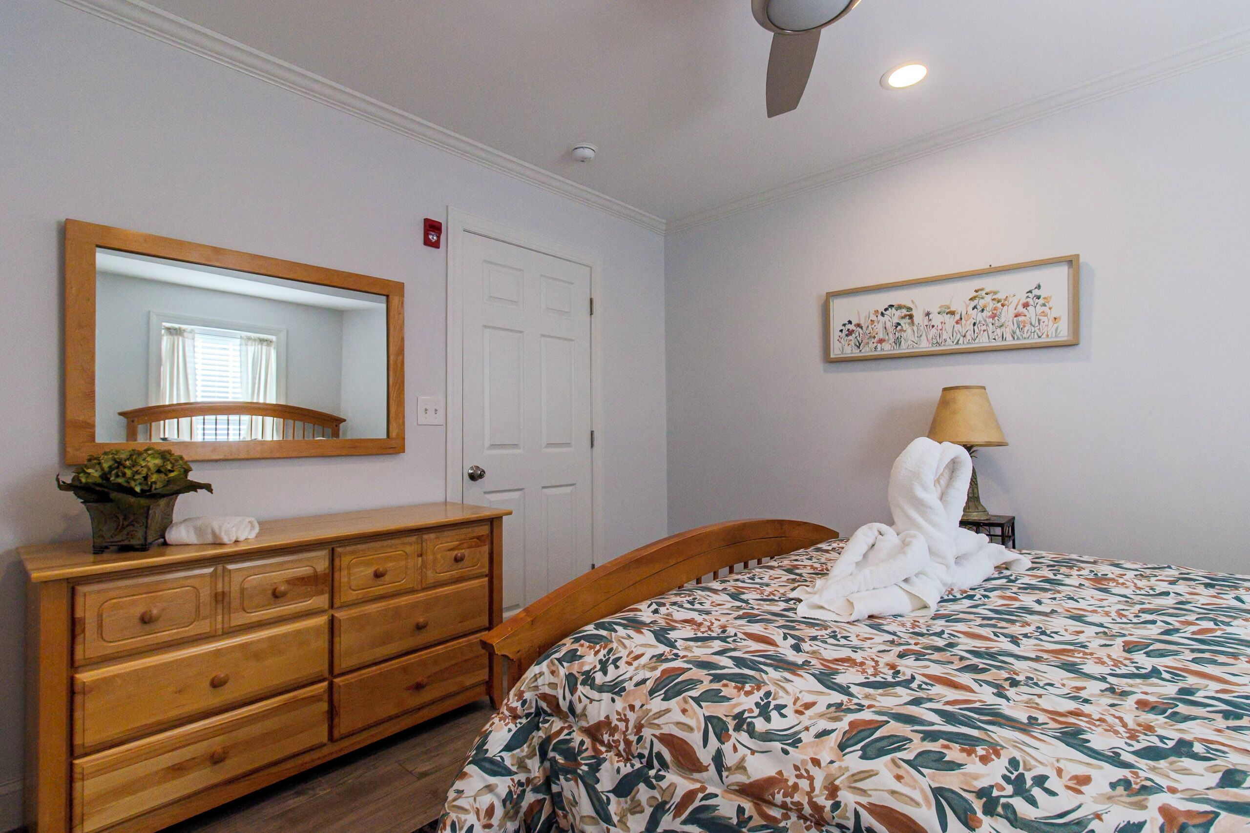 Um quarto com cômoda de madeira, cama com colcha florida, espelho, porta e luminária na parede.