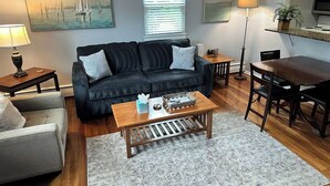 A living room with a dark grey sofa, a wooden coffee table, a grey armchair, a dining table with chairs, and a large window with blinds. - Comfy 2 BR Mid Term Apartment (Narberth)