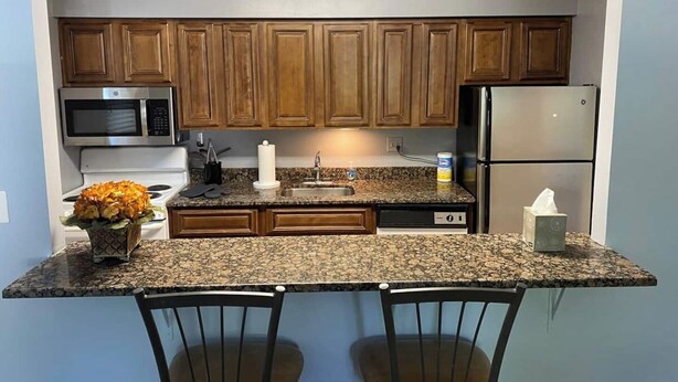 A compact kitchen with wooden cabinets, a granite countertop, a microwave, a refrigerator, and two chairs. - Comfy 2 BR Mid Term Apartment (Narberth)
