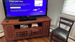 A television on a wooden stand with a Roku interface, a chair, and a window with blinds. - Comfy 2 BR Mid Term Apartment (Narberth)