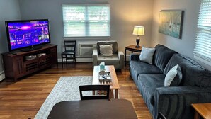 A living room with a flat-screen TV, a grey sofa, a wooden coffee table, a small table with a lamp, and a window with blinds. - Comfy 2 BR Mid Term Apartment (Narberth)