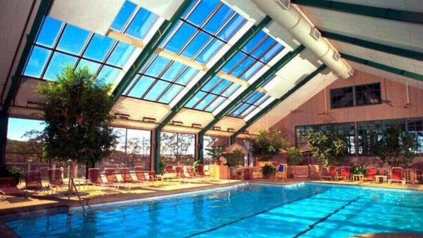 En indendørs swimmingpool med glaslofter, røde liggestole og udsigt over det omkringliggende landskab.