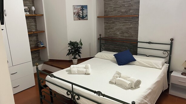 Un lit bien fait, avec du linge de lit blanc et un coussin bleu, une plante en pot et un mur de pierres en arrière-plan.