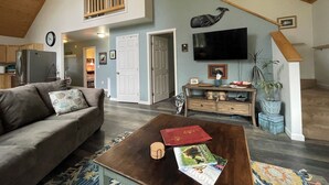 A spacious living area with a high ceiling, a large window, a flat-screen TV mounted on the wall, a sofa, a coffee table with books, and a staircase leading to an upper level. - Cranes' Rest: Cozy Homer Family Home Near Downtown & the Spit (Homer)