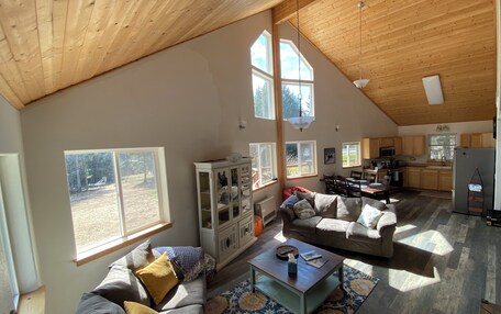 A spacious living area with a high ceiling, large windows, a dining table, and a kitchen in the background.