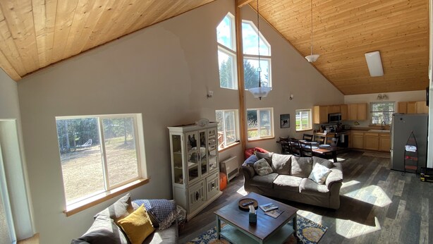 A spacious living area with a high ceiling, large windows, a dining table, and a kitchen in the background. - Cranes' Rest: Cozy Homer Family Home Near Downtown & the Spit (Homer)