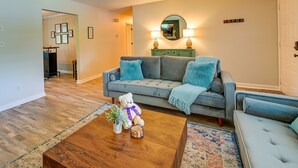 Un salon avec un canapé gris, une table basse en bois et un ours en peluche. Il y a un miroir au mur et une porte à l’arrière-plan.