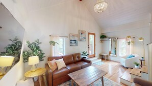 Ein geräumiges Wohnzimmer mit einem braunen Ledersofa, einem Holztisch und einem Kronleuchter. Es gibt Blumentöpfe, einen Spiegel und Kunstwerke an den Wänden.