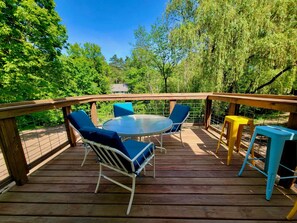 A wooden deck with a glass table and blue and yellow chairs, surrounded by lush greenery. - Colby Cottage in Taylors Falls, 4 Bedroom Home - Walk to Interstate Park (Taylors Falls)