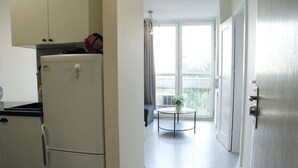 A compact kitchen with a refrigerator, a small table with a plant, and a view of the outdoors through a large window. - Central New Flat. AC, Park View , 2 Rooms (Warszawa)
