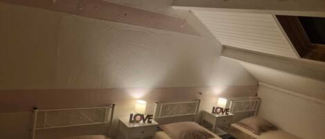A small, cozy room with two single beds, each with a nightstand and a lamp. The walls are painted in a light color, and there is a sloped ceiling.