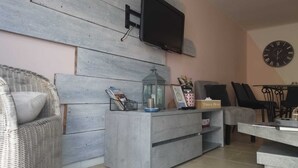 Una sala de estar con un sofá gris, un sillón de mimbre, una consola gris con un televisor, un reloj y una caja de pañuelos.