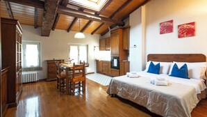 A hotel room with a wooden floor, a bed with white and blue bedding, a wooden headboard, a dining area with a table and chairs, and a kitchen area with wooden cabinets and a stove. - Residence Antico Borgo (Modena)
