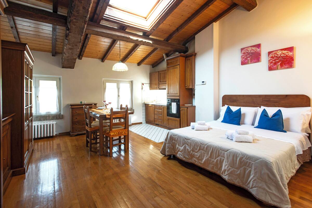 A hotel room with a wooden floor, a bed with white and blue bedding, a wooden headboard, a dining area with a table and chairs, and a kitchen area with wooden cabinets and a stove.