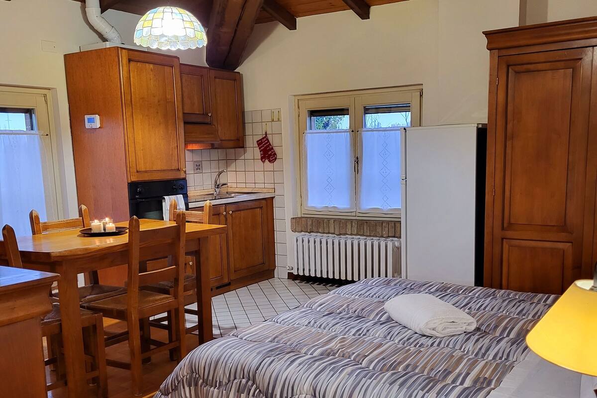 A cozy room with a wooden ceiling, a bed with striped bedding, a wooden dining table with chairs, a kitchen area with wooden cabinets, and a window with curtains.