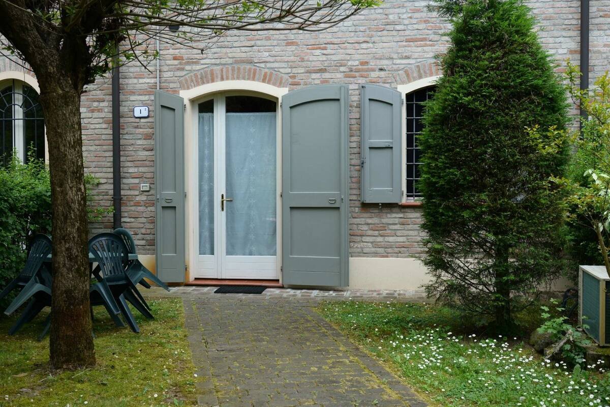 A brick building with a central door, two windows with shutters, and a small garden area with a tree and flowers.