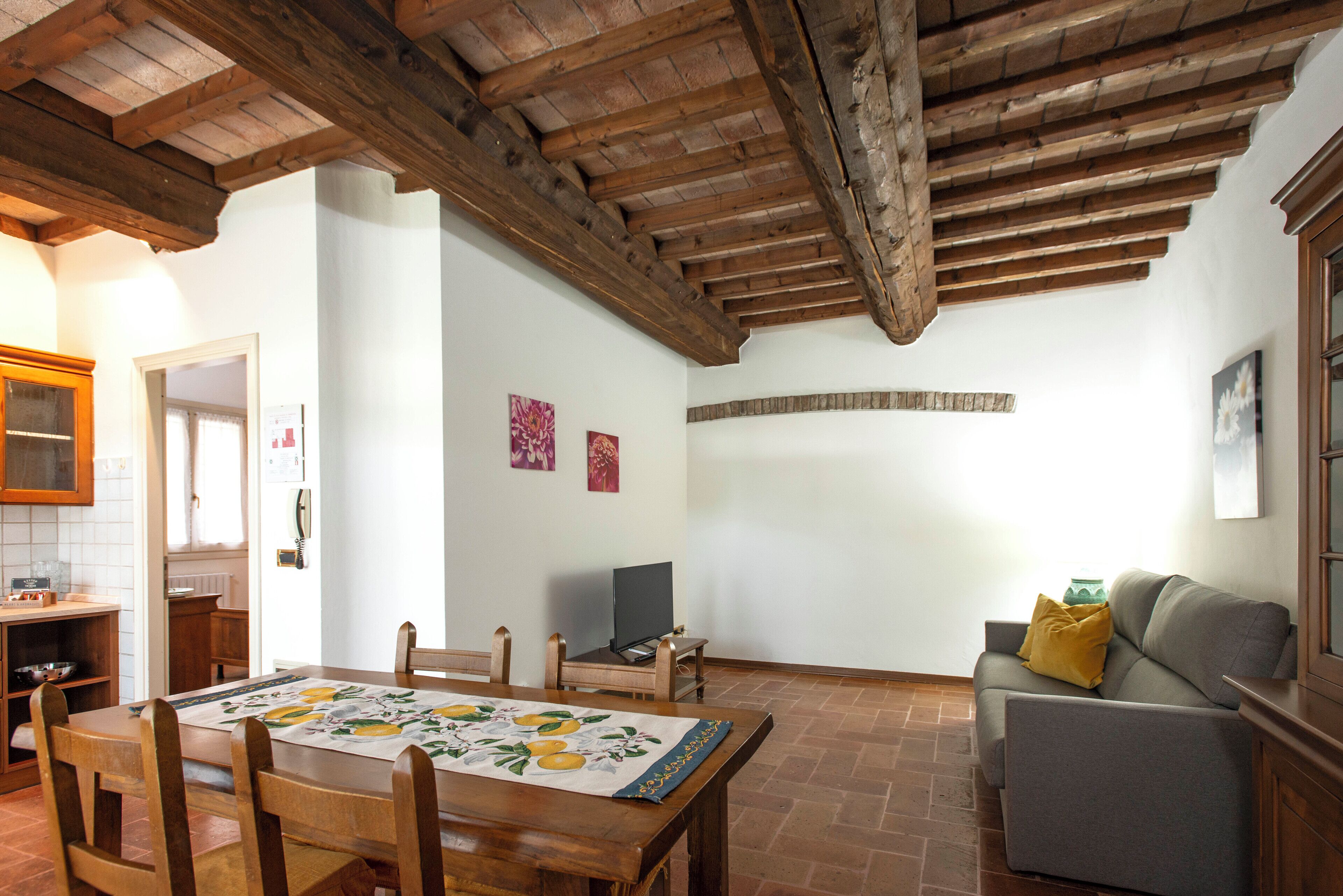 A dining area with a wooden table and chairs, a sofa, and a television in a room with exposed wooden beams and tiled flooring.