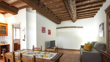 A dining area with a wooden table and chairs, a sofa, and a television in a room with exposed wooden beams and tiled flooring.