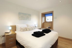 A neatly made bed with white linens and two black towels, a bedside lamp, a nightstand with a lamp, a painting on the wall, and a window with a blue blind.