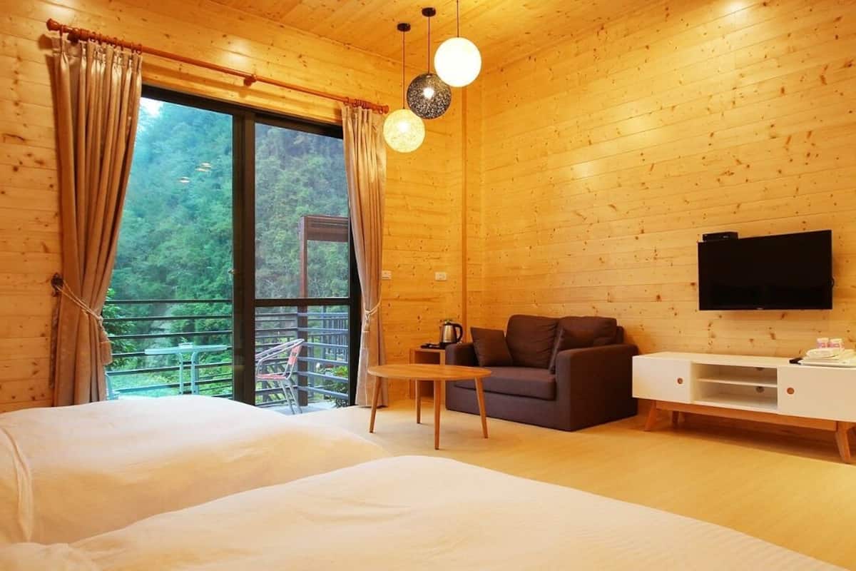 A wooden cabin room with a bed, a sofa, a TV, and a balcony with a view of greenery.